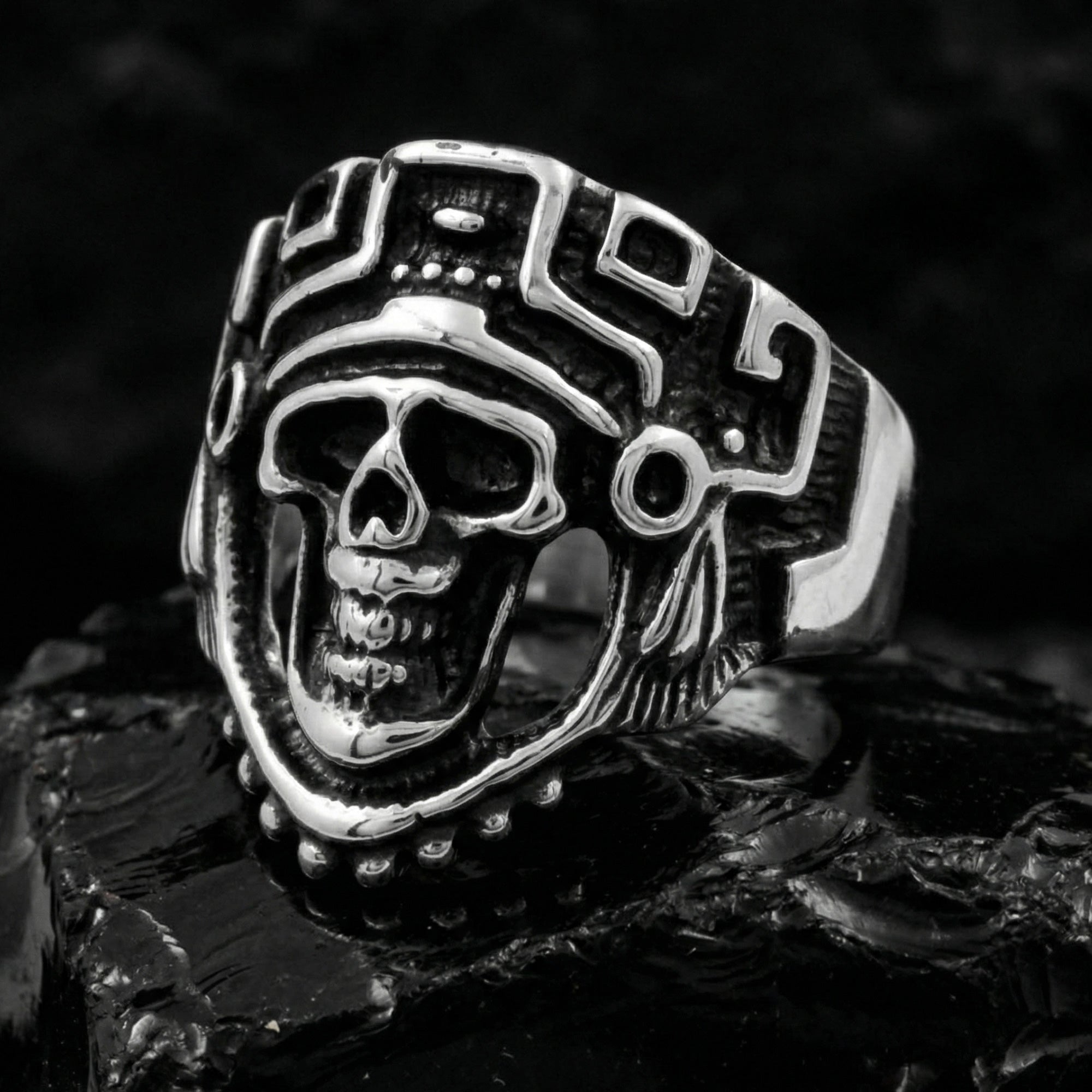 Stainless steel men's skull Aztec death mask ring sitting on black molten obsidian rock.