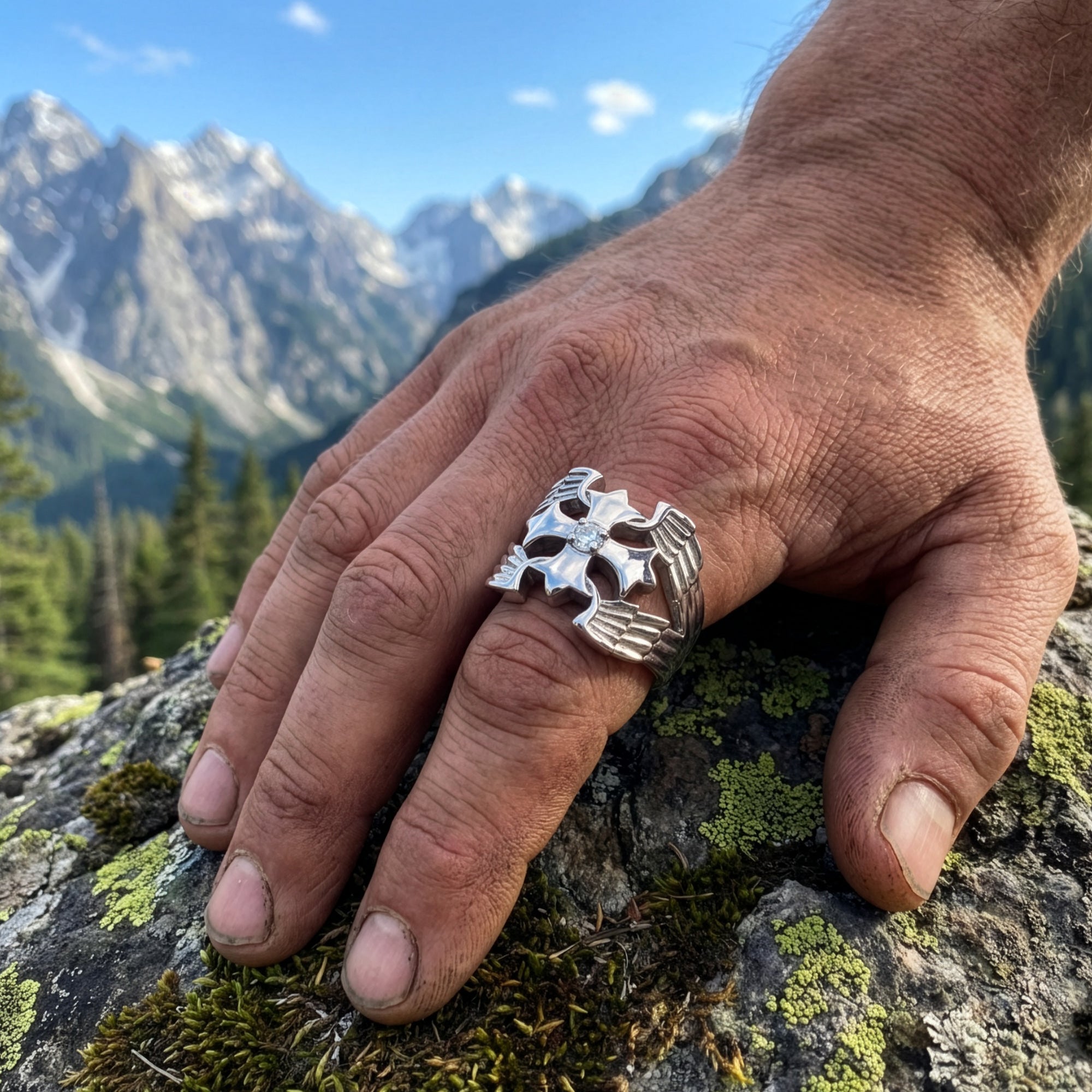 A man's hand with a winged cross stainless steel ring resting on a moss-covered rock with mountains in the background.