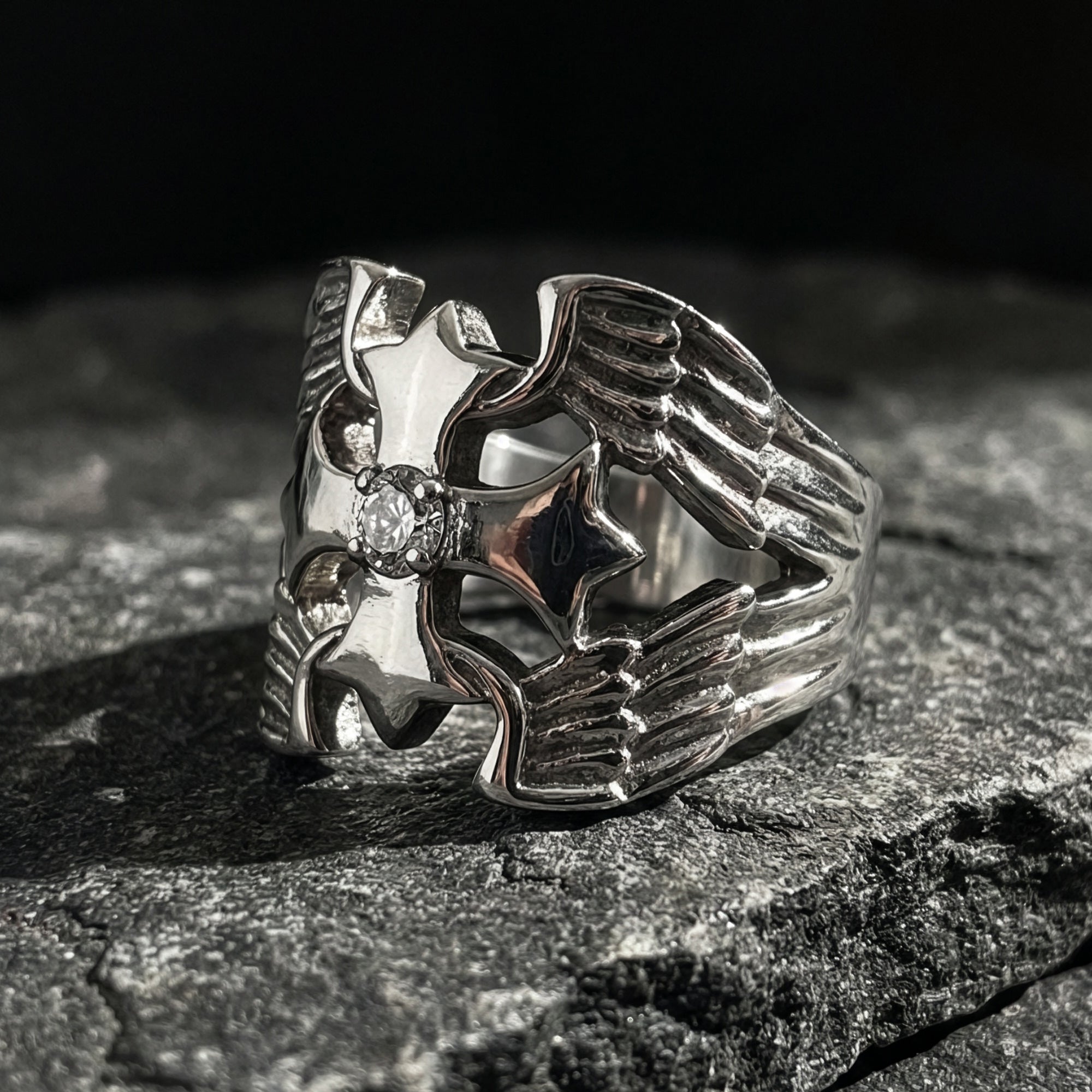A winged barbed cross stainless steel men's ring sitting on a slab of dark grey stone,