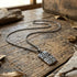 Black steel men's cross dog tag necklace laying on a wooden workshop bench.