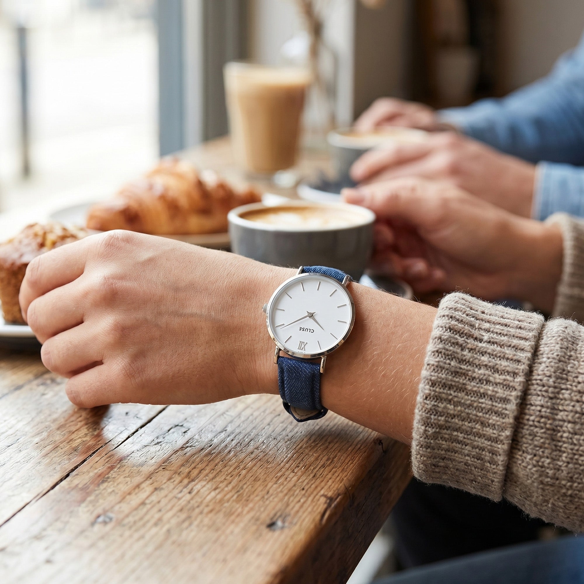 Woman wearing Cluse Minuit silver/white denim watch at an indoor cafe.