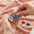 A woman places an Engelsrufer air element Luna soundball into a sterling silver carriage pendant. The three other soundballs in the collection lay on a pink fabric below.