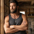 Man in a singlet wearing a fleur-de-lis dog tag necklace leaning against a timber cabin wall.