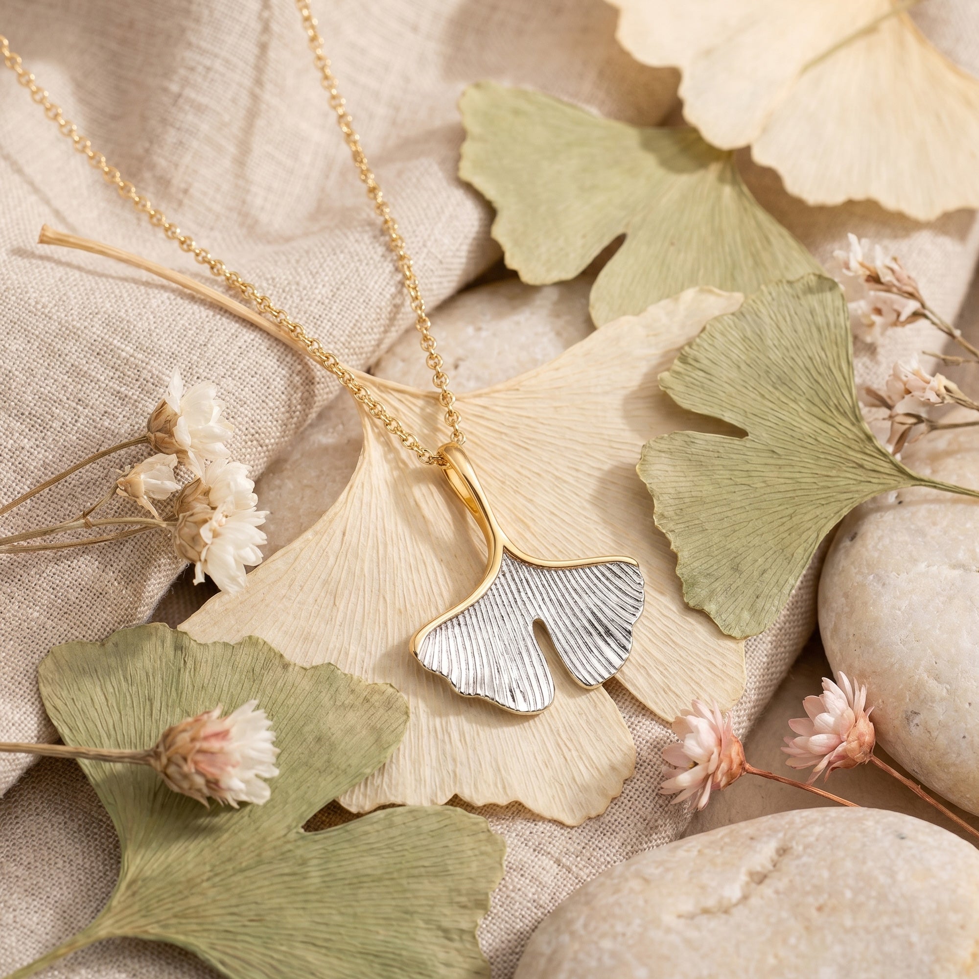 Engelsrufer ginkgo leaf gold silver necklace laying on a linen cloth surrounded by dried ginkgo leaves.