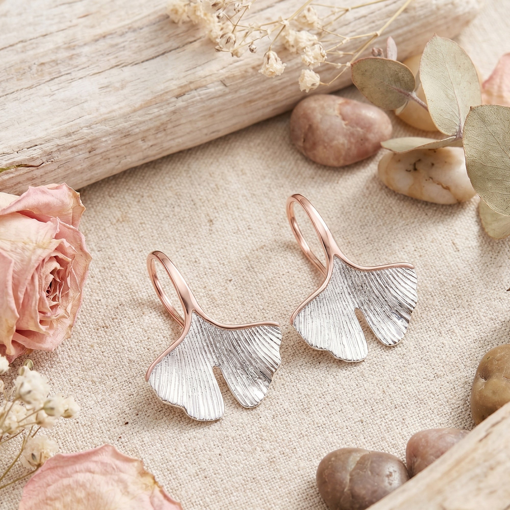 Engelsrufer gold ginkgo leaf hook earrings laying on linen cloth surrounded by a soft colour palette of dried flowers, wood and pebbles.