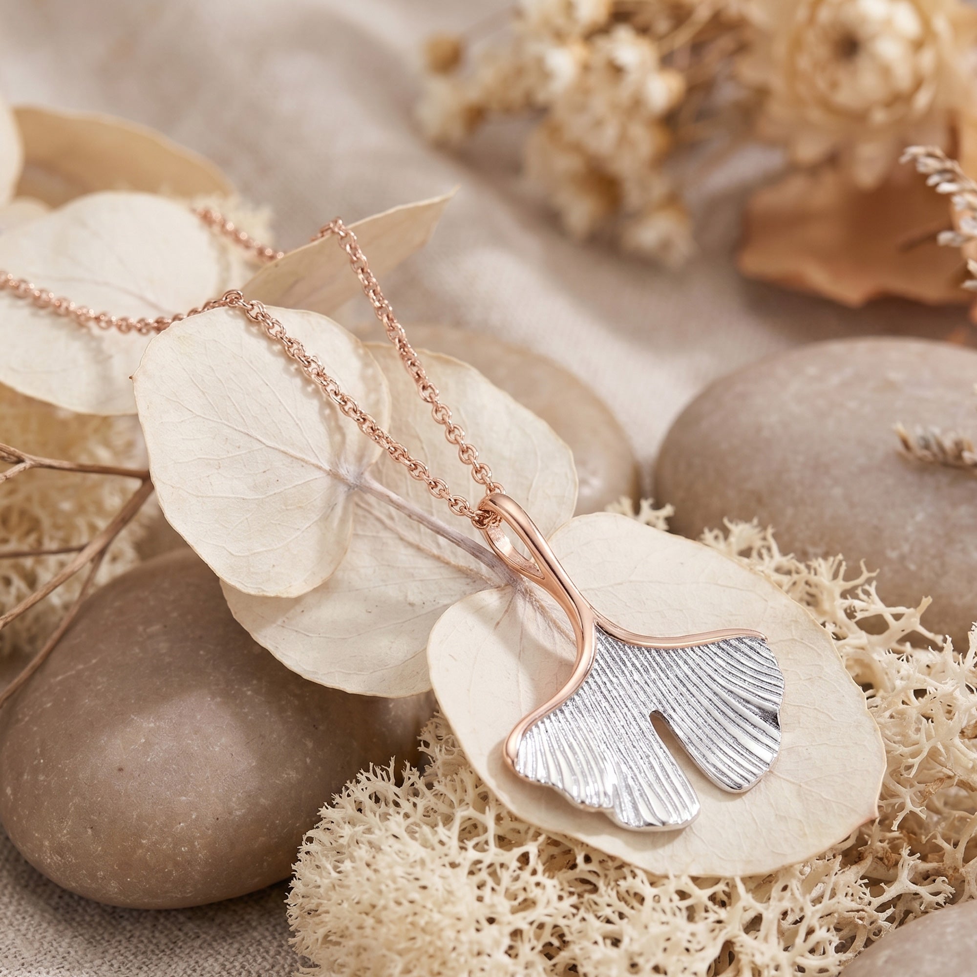 Engelsrufer ginkgo leaf rose gold silver necklace laying on dried leaves and small pebbles.
