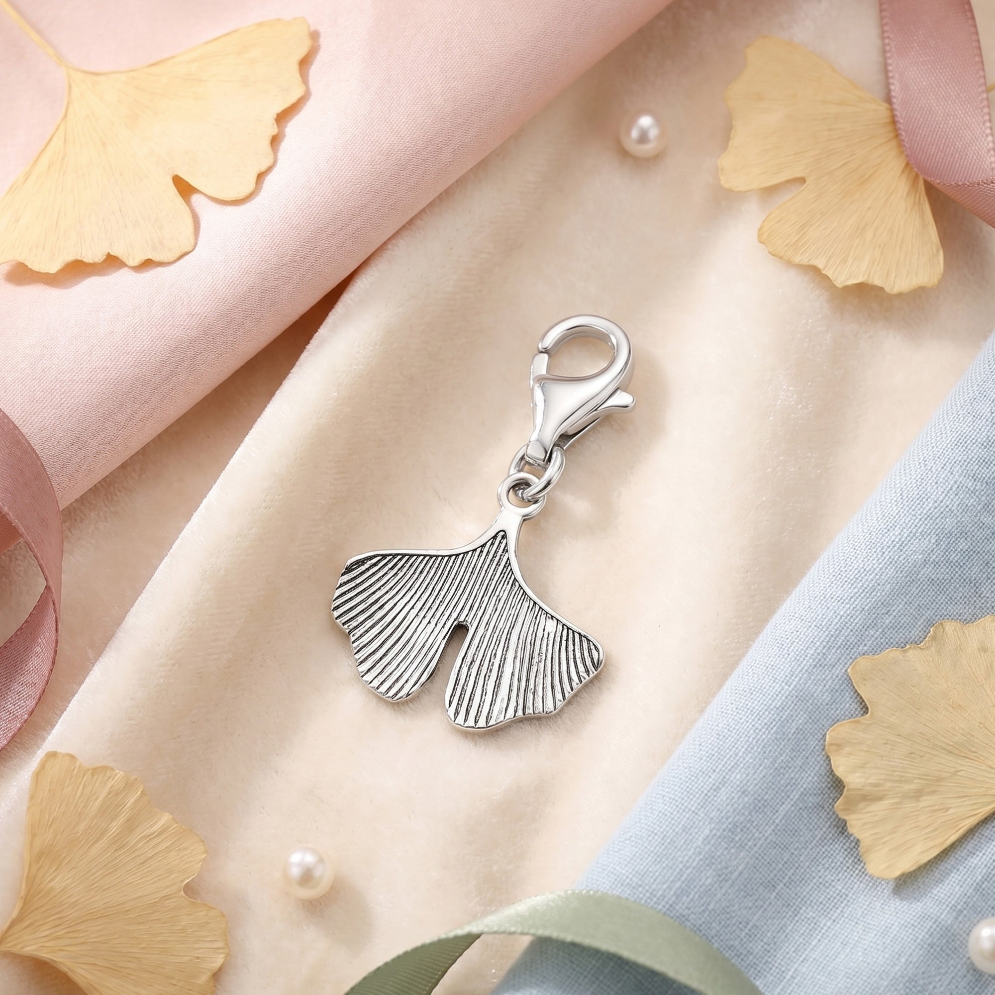 Engelsrufer ginkgo silver charm laying on a soft pallet of fabrics surrounded by dried ginkgo leaves.