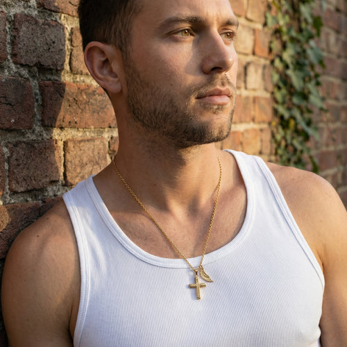 Man leaning against a brick wall wearing faith cross & wing steel gold necklace.