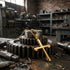 Save Brave barbed cross men's gold steel necklace laying against metal cogs in an industrial workshop.