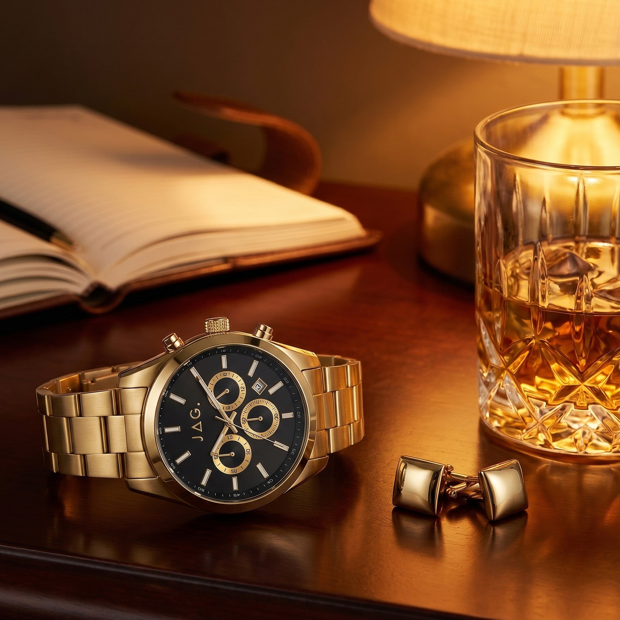 JAG Acton men's black dial gold watch on a wooden desk with a journal, cufflinks and a whisky glass.