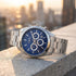 JAG Acton men's blue dial silver watch on a concrete ledge with a skyline of buildings in the background.