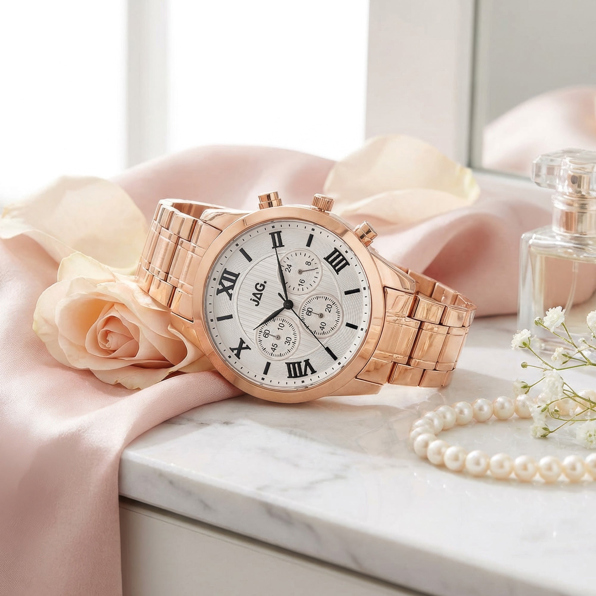 JAG ladies Estelle rose gold watch laying on white marble dressing table with pink fabric, pearls and perfume.