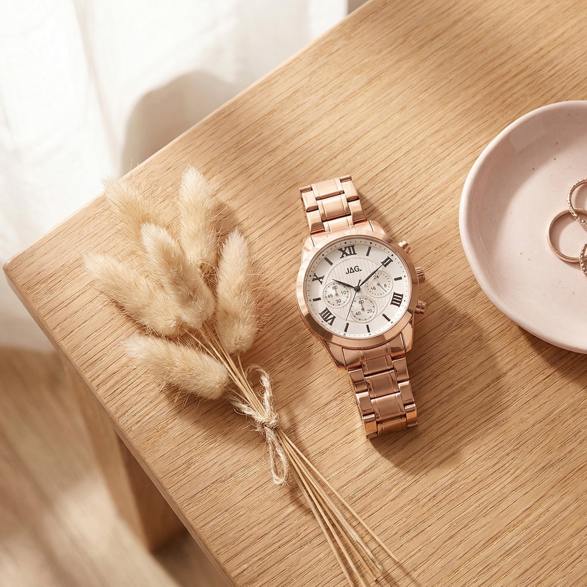 JAG ladies Estelle rose gold watch laying on a wooden table with dried flowers and jewellery.