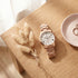 JAG ladies Estelle rose gold watch laying on a wooden table with dried flowers and jewellery.