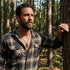 Man in an open-neck shirt leaning against a tree in the forest wearing military star dog tag necklace.
