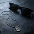 Police Men In Black stainless steel dog tag limited edition necklace on leather surface with black pair of sunglasses.