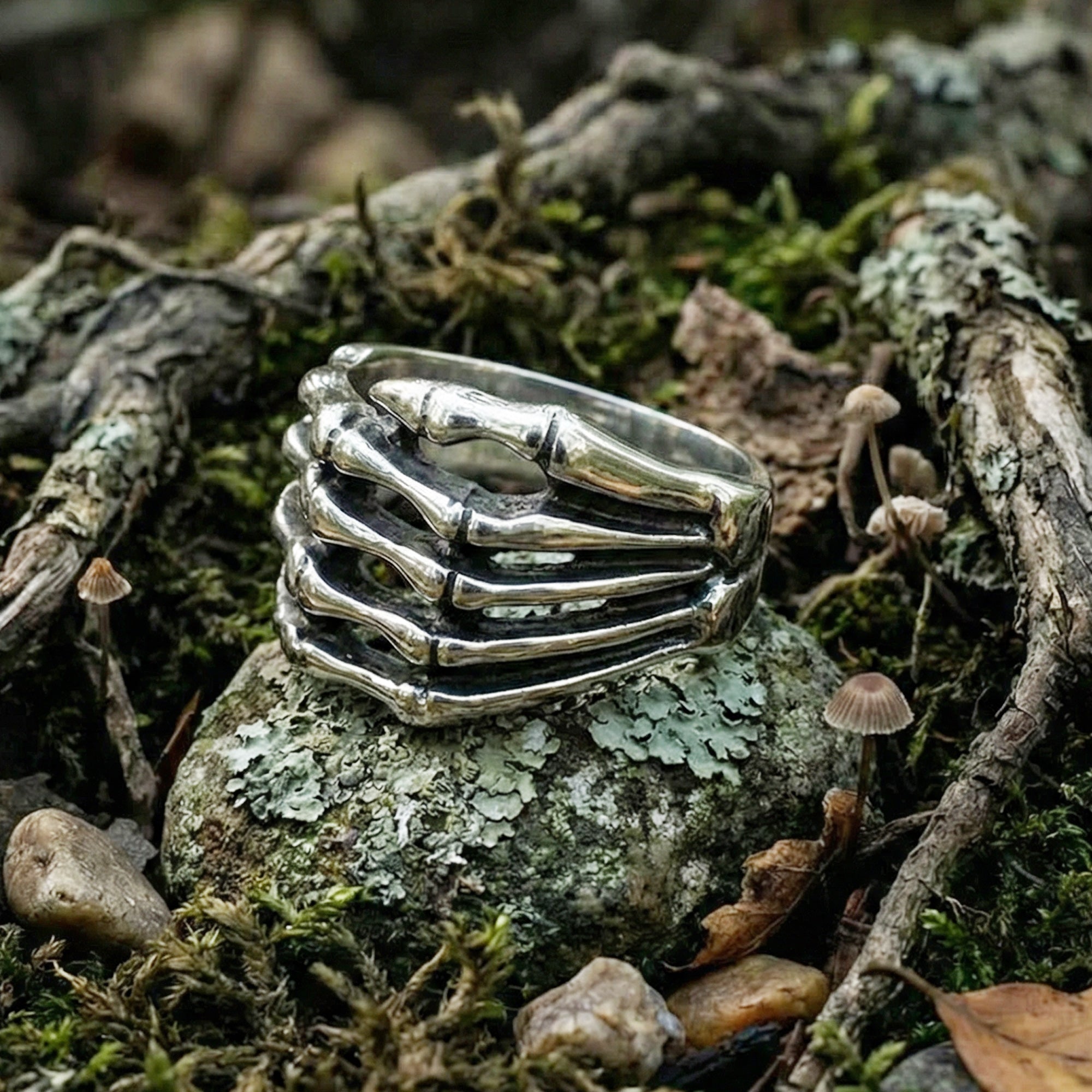 Stainless steel men's skeleton hand ring resting on a moss-covered rock surrounded by tree roots.
