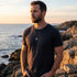 Man standing on rocks near the foreshore wearing an Anchor & Crew Union anchor sterling silver necklace.