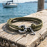 Anchor & Crew Clyde sterling silver khaki green rope bracelet on wooden jetty with barnacles and a boat in the background.
