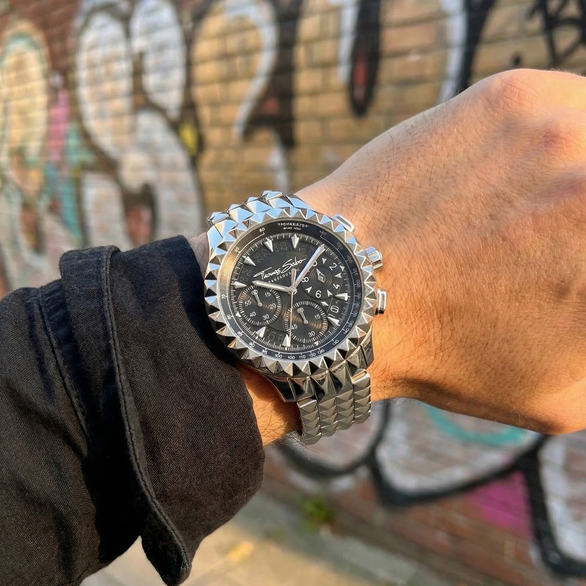 Man wearing Thomas Sabo Rebel At Heart silver pyramid men's watch with a graffiti covered wall in the background.