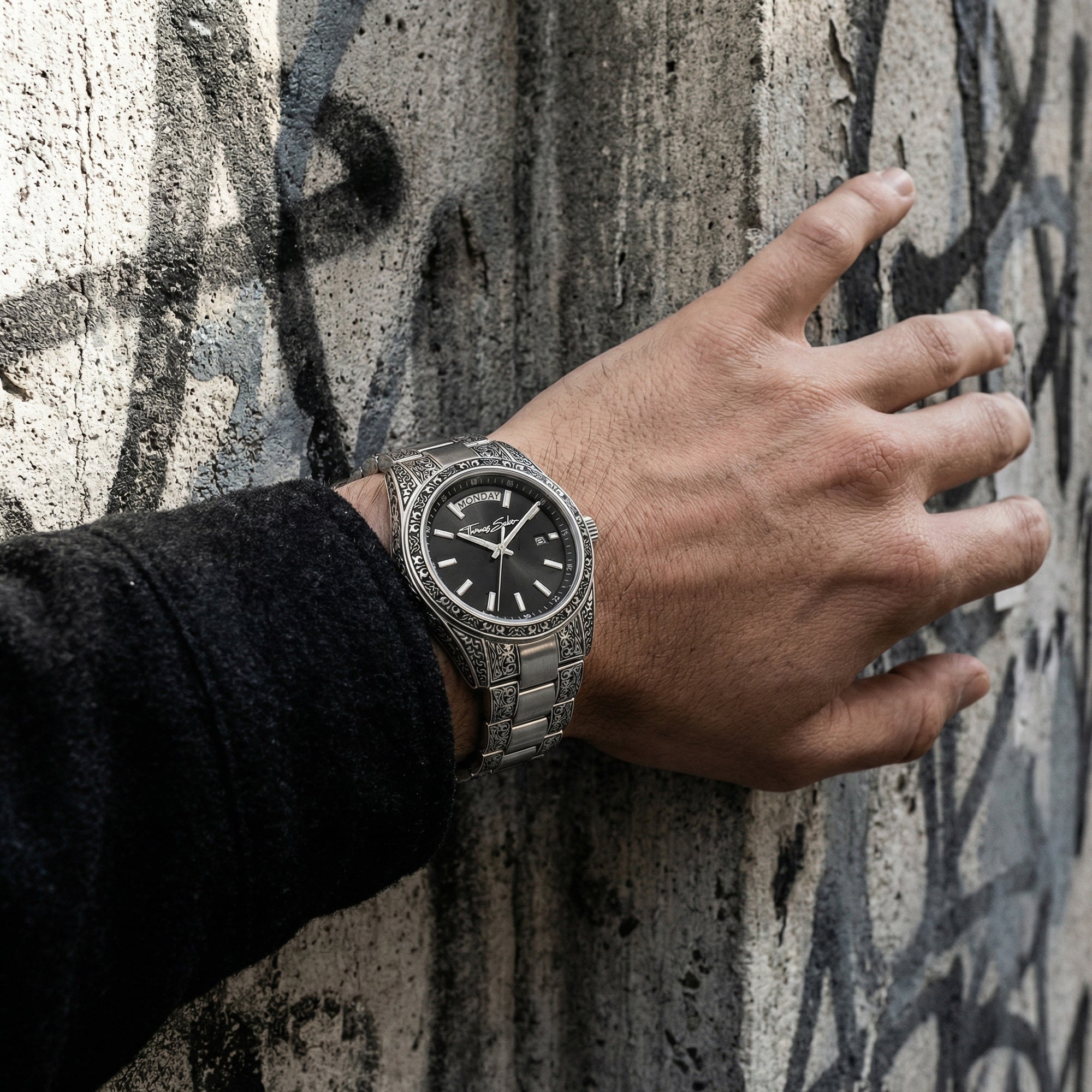 Man wearing a Thomas Sabo Rebel At Heart Revive 36mm black dial watch resin this hand on a concrete wall with graffiti. 