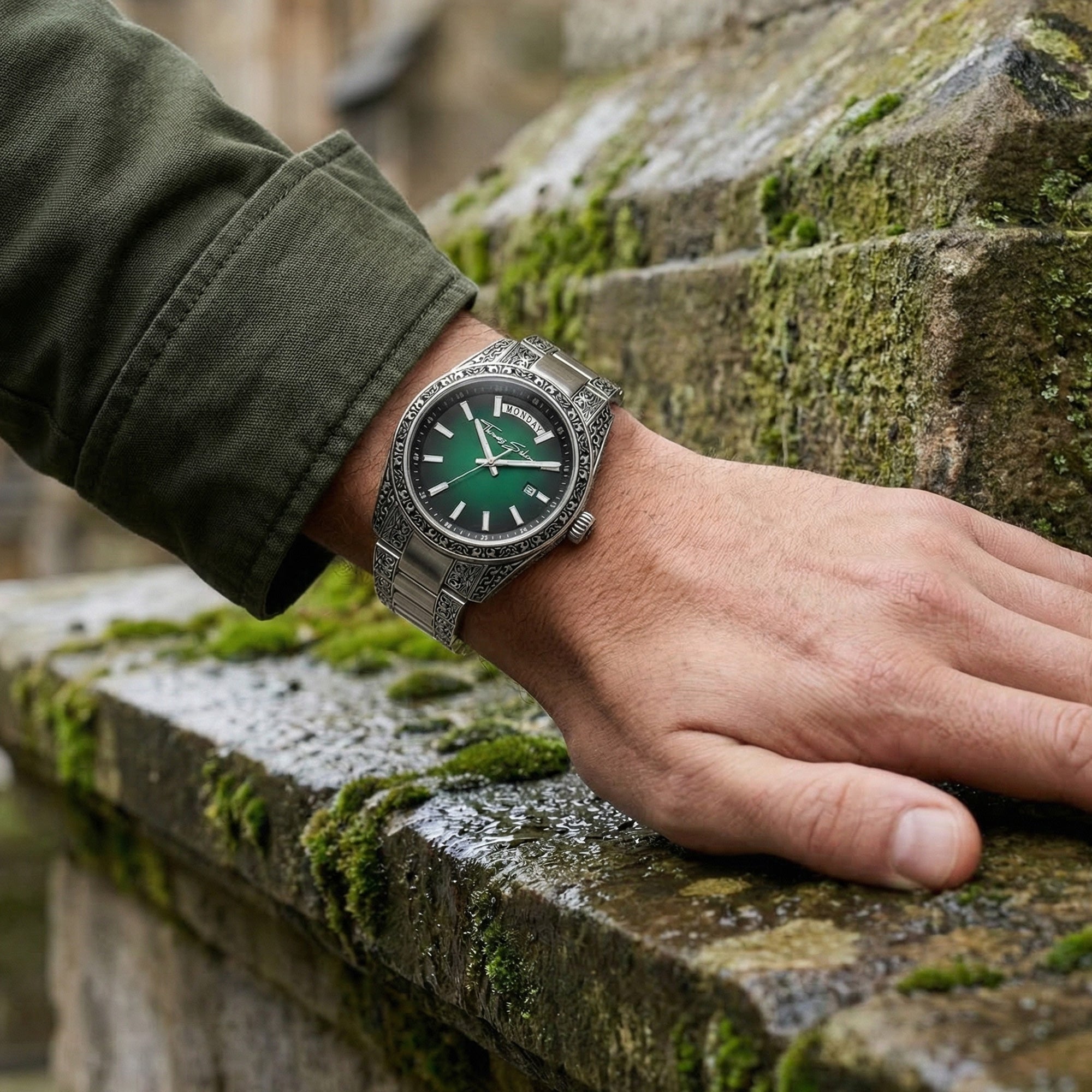 Man wearing a Thomas Sabo Rebel At Heart Revive 40mm green dial watch wresting his hand on the moss-covered block work of an old building.