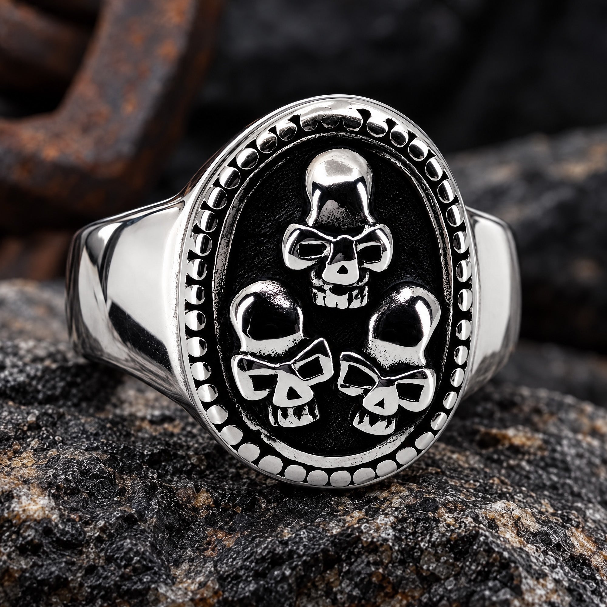 A three skulls stainless steel men's signet ring sitting on a broken rock with rusted iron remains in the background.