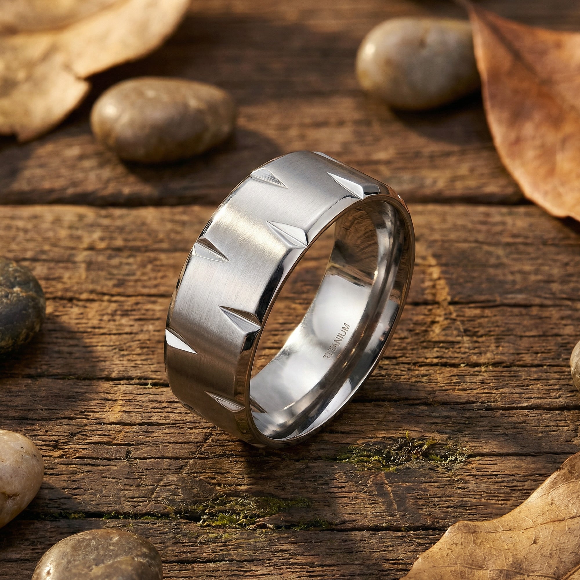 Titanium men's matte finish notched edge ring on a wooden surface surrounded by small rocks and leaves.