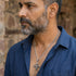 Close up of a bearded man in a blue open-neck shirt looking to the left while wearing a vajra cross sterling silver necklace.