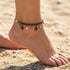 Close-up of a woman's foot in the sand wearing an Engelsrufer wind rose gold and black boho pearl anklet.