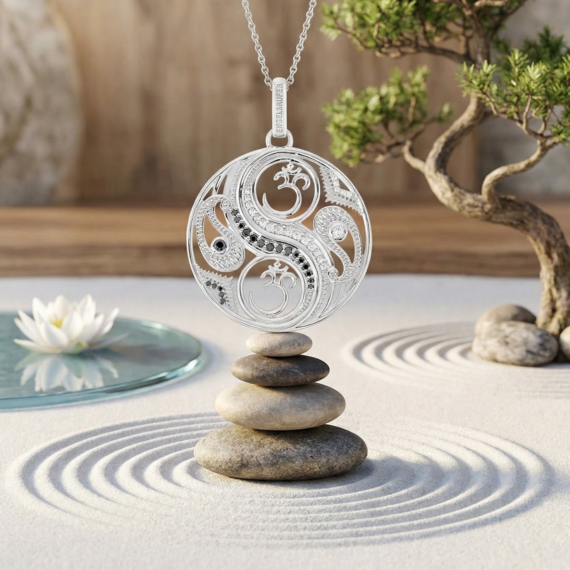 An Engelsrufer yin yang om balance pendant suspended over a stack of balanced stones on sand with a bonsai tree in the background.