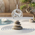 An Engelsrufer yin yang om balance pendant suspended over a stack of balanced stones on sand with a bonsai tree in the background.